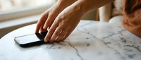 Woman's hand reaching for smartphone on marble table surface near window with natural lighting for modern lifestyle and technology concepts.