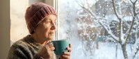 Senior Caucasian woman in knit hat enjoying hot beverage by window during winter snowfall, cozy indoor moment with peaceful expression and warm atmosphere.