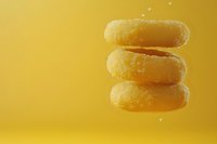 Stack of golden glazed donuts floating in air against vibrant yellow background, with subtle crumbs and particles creating dynamic food composition.