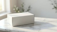 White cardboard box on marble surface with natural lighting and green plants in background for product packaging mockup and branding presentation.