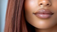 Close up view of plump glossy lips with natural nude pink lipstick and smooth dark skin texture showing part of nose and straight brown hair against blue background.