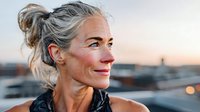 Confident mature woman with silver hair in messy bun gazing at sunset, urban rooftop setting, natural makeup, glowing skin, peaceful expression.