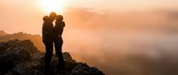 Romantic couple silhouette embracing on mountain peak during golden sunset with misty valleys below creating intimate moment for love and adventure themes.