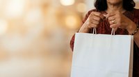 Woman with dark hair and medium skin tone holding white shopping bag against blurred warm background, retail and consumer lifestyle concept.