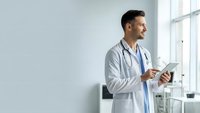 Young male doctor in white coat using digital tablet in modern hospital office, smiling while reviewing patient data for healthcare technology solutions.