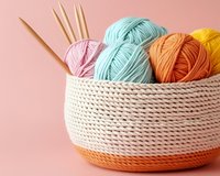 Colorful yarn balls in mint, orange, yellow and pink shades arranged in knitted basket with bamboo needles on pink background. Perfect for craft and hobby themes.