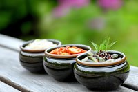 Korean side dishes in traditional stone bowls with kimchi, sesame seeds and fresh herbs on wooden surface against blurred garden background.