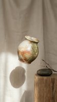 Rustic handmade ceramic vase with earthy glaze tones floating above a wooden stump with a stone and twig, soft natural light and linen backdrop.