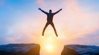 Silhouette of man jumping between mountain peaks at sunset, expressing freedom and achievement against dramatic sky with golden sunburst background.