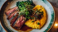 Grilled medium-rare steak slices with rich sauce, served alongside creamy yellow polenta and topped with fresh microgreens on rustic ceramic plate.