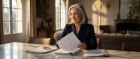 Mature Caucasian businesswoman reviewing financial documents at home office desk with warm natural lighting from windows creating professional workspace atmosphere.