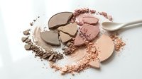 Broken makeup powder compacts with scattered cosmetic dust and brush on white background for beauty and fashion projects.