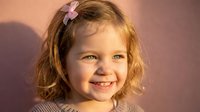 Happy young girl with blonde curly hair and pink bow smiling warmly in golden sunlight against soft pink background for family lifestyle content.
