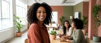 Young African American businesswoman smiling at camera in modern office with diverse female colleagues working at table in background.
