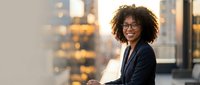 Young woman with curly dark hair and glasses wearing navy blazer, smiling at camera in modern office with city skyline background, professional business portrait.