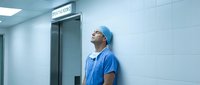 Exhausted male surgeon in blue scrubs leaning against hospital wall after long surgery, showing fatigue and stress in medical environment.