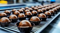 Rows of chocolate truffles in dark paper cups on production line conveyor belt, selective focus on glossy round confectionery desserts in factory setting.