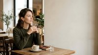 Young woman with dark hair and olive complexion, wearing olive green sweater, sitting at wooden table with coffee cup and glass of water, contemplative expression, modern interior setting with plants and natural light.