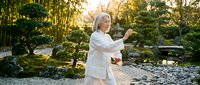 Senior woman with white hair practicing tai chi or qigong in a serene garden with water feature and lush greenery during golden hour, promoting wellness and mindfulness.