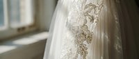 Elegant white wedding dress with intricate lace floral embroidery detail hanging by sunlit window in bridal preparation room for ceremony celebration.