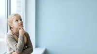 Young child with blonde curly hair, Caucasian ethnicity, wearing beige knit sweater, sitting by window in contemplative pose, hand to chin, looking upward thoughtfully in bright modern interior space.