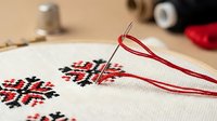 Cross stitch embroidery hoop with needle and red thread on linen fabric, featuring traditional folk ornament pattern in red and black.