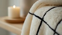 Soft white towels with black geometric pattern neatly folded on wooden shelf next to glowing candle creating relaxing spa atmosphere. Wellness and self-care concept.