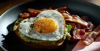 Sunny side up egg on avocado toast with crispy bacon strips on dark plate, seasoned with black pepper and herbs, morning breakfast close-up.