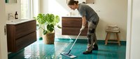Man in casual work clothes mopping turquoise tile floor in modern home interior with wooden furniture and potted plant, cleaning service and household maintenance.
