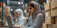 Young Middle Eastern woman in hijab and striped shirt working on laptop in warehouse setting with cardboard boxes and storage shelves in background.
