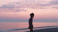 Woman silhouette standing on beach at sunset with pink purple sky reflecting on calm ocean water creating peaceful meditation moment for wellness relaxation.