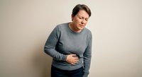 Middle-aged Caucasian woman experiencing stomach pain holding abdomen in gray sweater against neutral background for health and wellness concepts.