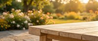 Empty wooden table surface with blurred garden background featuring blooming flowers and sunlight, suitable for product placement and outdoor lifestyle content.