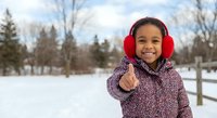 Happy mixed-race girl in winter coat and red earmuffs pointing at camera in snowy park with evergreen trees and fence background for seasonal advertising.