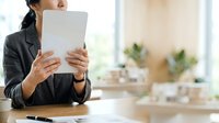 Young woman of East Asian descent in professional business attire holding tablet device at desk, suitable for corporate, technology, and workplace concepts.