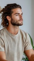 Young Caucasian man with beard and long wavy hair in casual beige t-shirt looking thoughtfully to side against light background, profile portrait.
