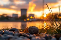 Tranquil riverside scene at golden hour with stones and grass silhouettes against industrial cooling towers, dramatic sunset sky reflecting on calm water surface.