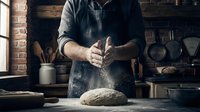 Baker dusting flour on fresh dough while working in rustic kitchen workshop. Male artisan, Caucasian, middle-aged, wearing apron, preparing bread for baking.