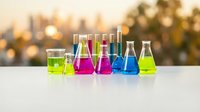 Colorful laboratory glassware with bright chemical solutions arranged on white surface against blurred city skyline at sunset for science education.