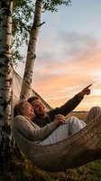 Happy couple relaxing in hammock between birch trees during golden hour sunset, pointing at sky while enjoying romantic outdoor moment together.