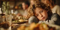 Sad young boy with curly hair resting head on arms at holiday dinner table, looking pensive while family celebrates in background.