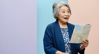 Senior Asian woman with gray hair holding and reading a map against colorful pastel background, planning travel or vacation.