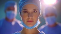 Female medical professional in surgical attire with protective mask lowered, looking confident against blurred background of operating room team and surgical lights.