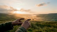 Hands framing golden sunset over rolling green hills creating natural viewfinder gesture during peaceful evening in countryside landscape.