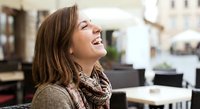 Happy young woman laughing outdoors at cafe terrace wearing brown scarf and sweater enjoying sunny day lifestyle moment for wellness and joy concepts.