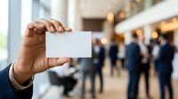 Close-up of male hand holding blank white business card in modern office lobby with blurred colleagues networking in background for corporate branding.