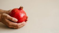 Hands holding fresh red pomegranate against neutral background, symbolizing health, nutrition, and organic fruit for wellness and food industry use.