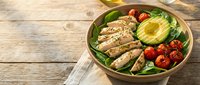 Grilled chicken breast slices with fresh avocado, cherry tomatoes and mixed greens in a bowl on wooden table for healthy lunch meal prep.