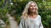 Mature blonde woman smiling outdoors in casual gray sweater with natural green foliage background for lifestyle and wellness content.