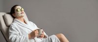 Young woman relaxing in spa chair wearing white bathrobe and eye mask, holding tea cup during wellness treatment session for self-care and beauty routine.